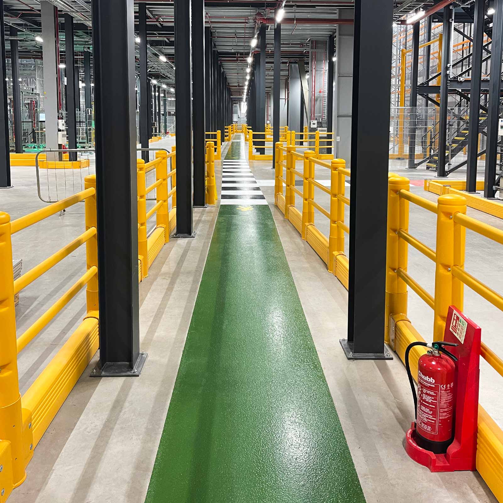 Green pedestrian walkway lined on both sides by SafeStop Pedestrian with GroundBarrier between steel columns in a warehouse.